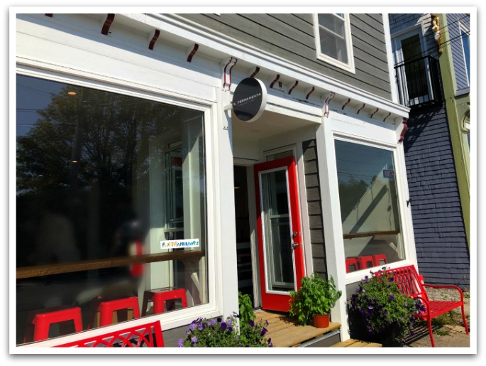 Exterior of Il Ferramenta showing seating by the windows, red bar stools, a red door, and red benches outside.