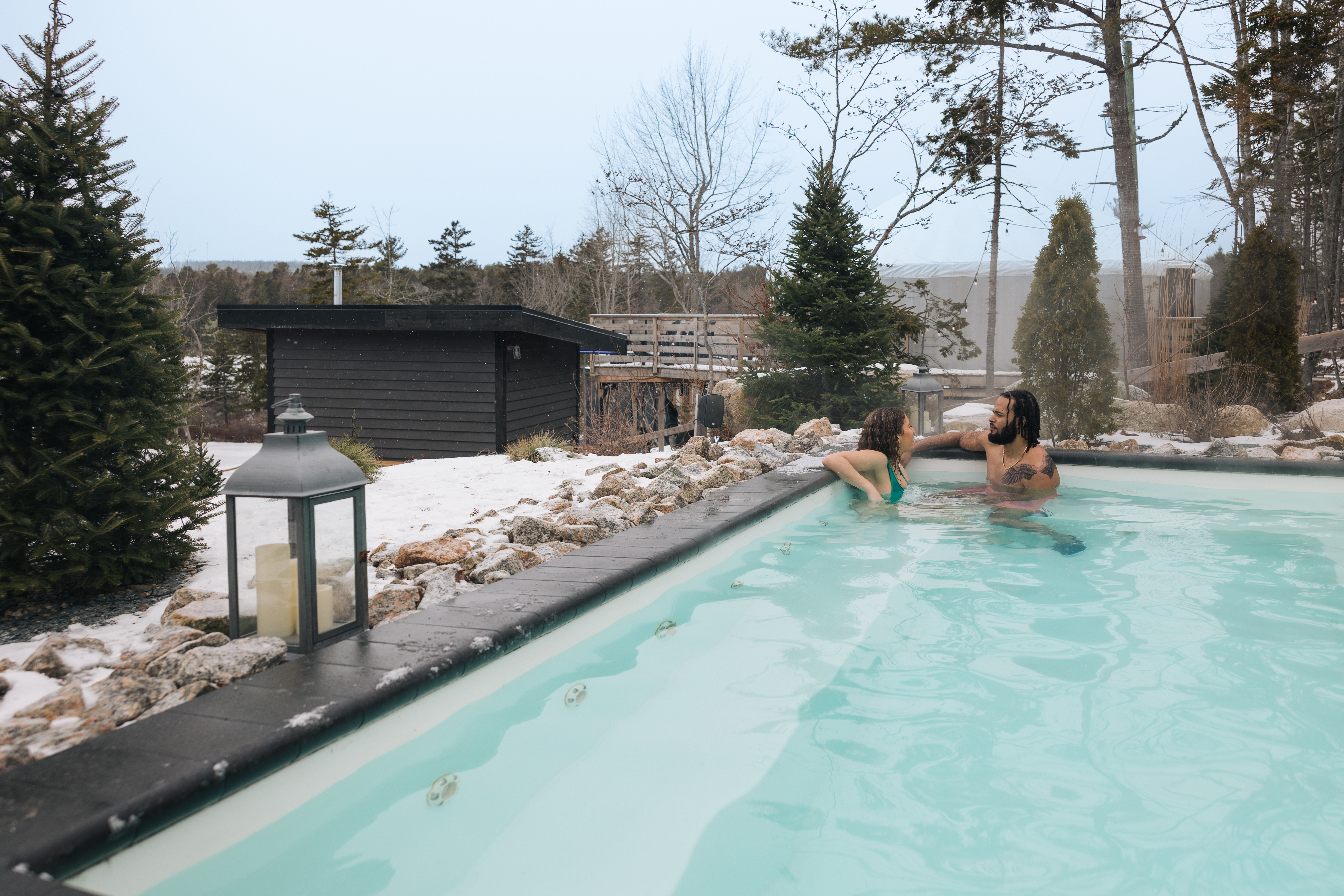 Couple lounging in the corner of a pool in winter with a small black building in the background.